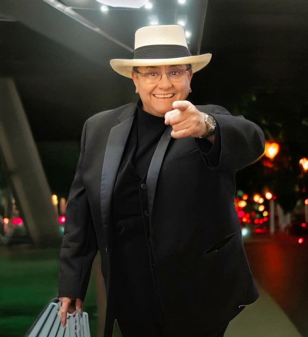 Photo of woman in all black suit and hat, standing and pointing towards the camera with a smile on her face.