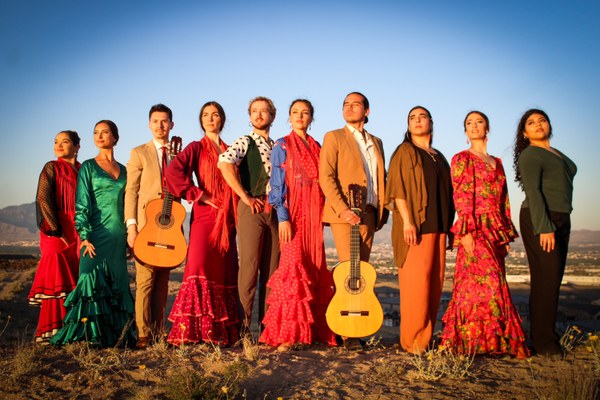 Photo of a line of 10 men and women standing, looking off past the camera. They are each in their flamenco dressed and suits. Two of them are holding guitars.