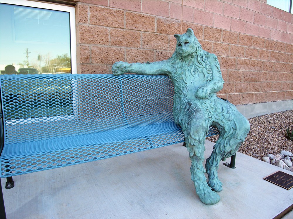 Bronze sculpture depicting a cat with one paw on the back of a bench. 