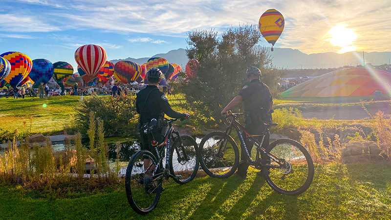 Two APD officers on bikes taking a rest on a grassy field with hot air balloons in the background.