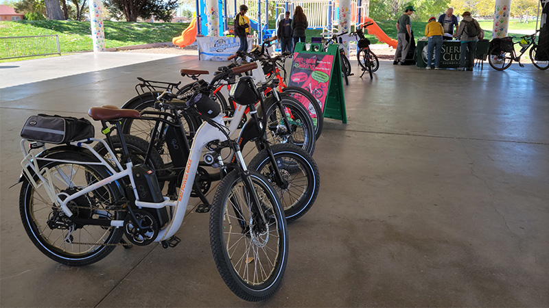 A row of electric bikes on a concrete floor of a covered, open building in a park.