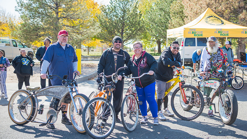 A group of adults pose and smile with the audience. All are either holding or sitting on their bikes. There are a mix of street and dirt bikes. Behind them is a park with lush green grass and trees. A One Albuquerque Esperanza yellow tent appears in the background.