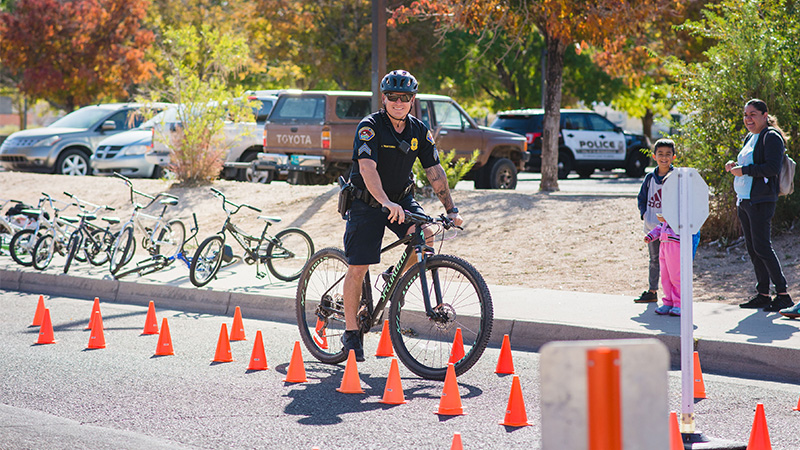 An Albuquerque Police officer in uniform and a helmet smiles at the audience while riding a bike through a bike course in a parking lot lined with small orange cones. Two children and an adult look on and smile from the sidewalk. A park full of trees and parked cars appear in the background.