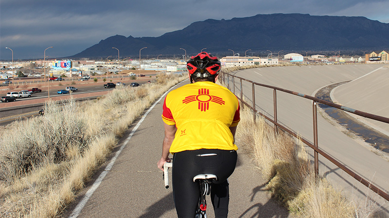 A cyclist bikes down a paved path next to an arroyo. He is wearing a helmet and a yellow shirt with a red Zia symbol on the back. The Sandia mountains appear in the background. Cars can be seen driving down the interstate in the bottom left hand corner.