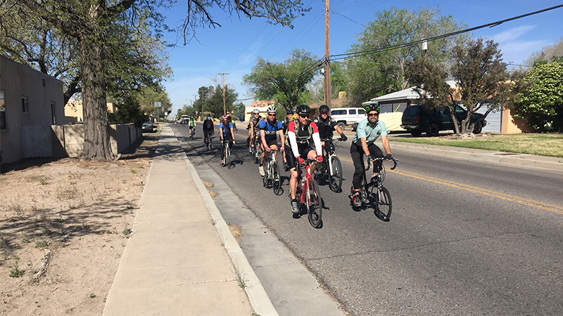 A group of cyclists riding two-by-two down a residential street in Albuquerque on a bright, sunny day. Trees, houses, and cars dot the road on either side. All cyclists are wearing helmets.