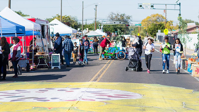 A group of cyclists and walkers talk and mingle during a CiQlovia event. Some folks are walking while others are visiting covered boths and speaking with various vendors.