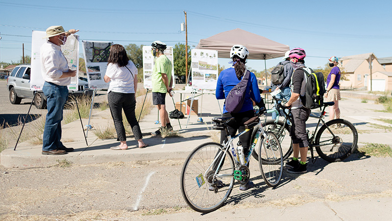 A small group of people standing and holding bikes review and discuss a series of posters with maps and diagrams on them in a parking lot near a residential area.