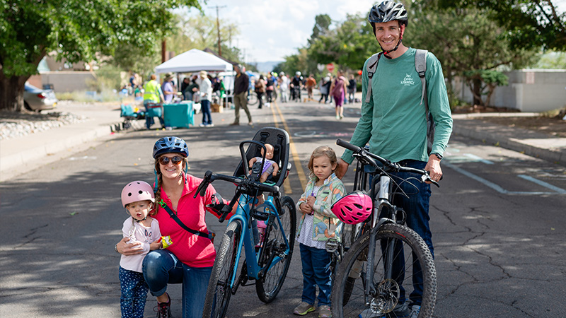 A family with two kids pose and smile at the audience. The mother is kneeling and hugging a small girl with one hand stabilizing her bike. The dad is standing with one hand on his bike and the other on his older daughter's back.