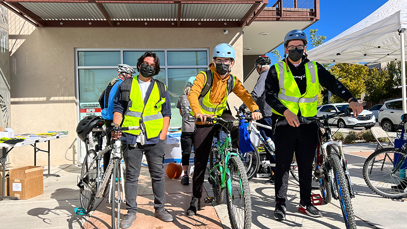 Three people stand on or beside their bikes and look at the audience. Behind them is a light tan, two-story adobe building.