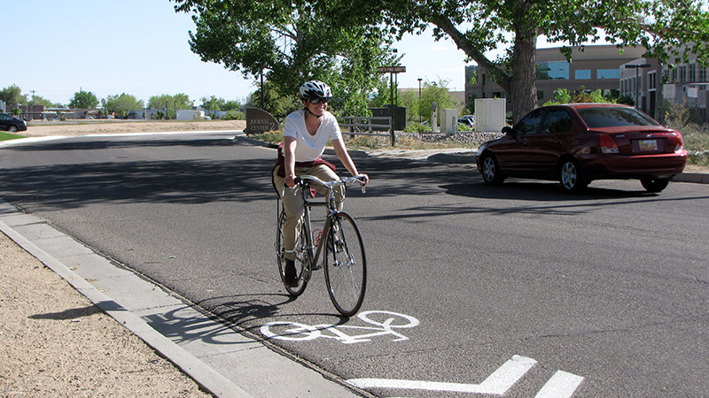 A woman wearing a helmet rides her bike in a painted bike lane section of a paved road next to a car. Trees are visible in the background.