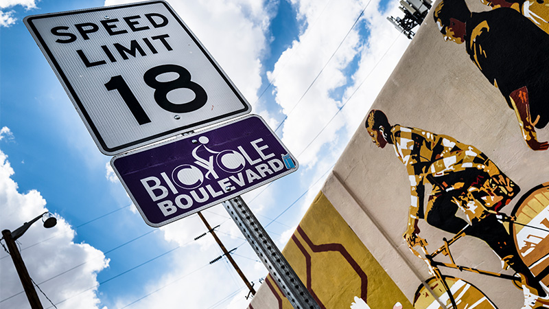 A close up of a speed limit sign (18 mph) with a plaque underneath that says "Bicycle Boulevard". Behind the sign is a mural of people riding bikes.