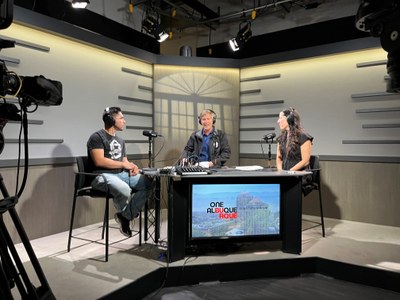 One ABQ & You Podcast Hosts Chatting Mayor Tim Keller, seen center, and Leah Black, seen right, and a podcast guest, seen left, sit at a triangular black table. Mayor Keller is speaking and the other two individuals are listening. All are wearing black headphones and have microphones in front of them. Small portions of the studio camera equipment can be seen in the edges of the frame on the left and right.