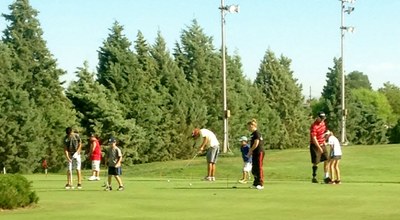 Puerto del Sol Junior Golf Photo A group of children and adults playing golf on a green field surrounded by pine trees.