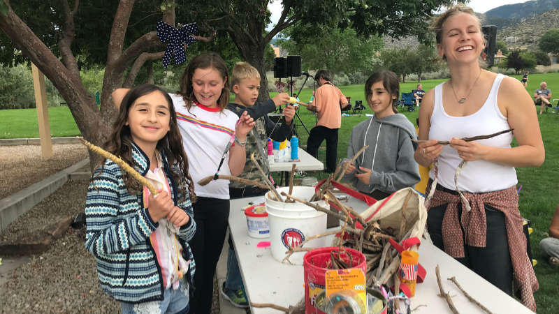 A group of kids making crafts from sticks at a table in a park.