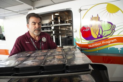 Menu Nutrition Home Tile A man holding a tray of prepackaged meals in front of a Nutrition Program truck.